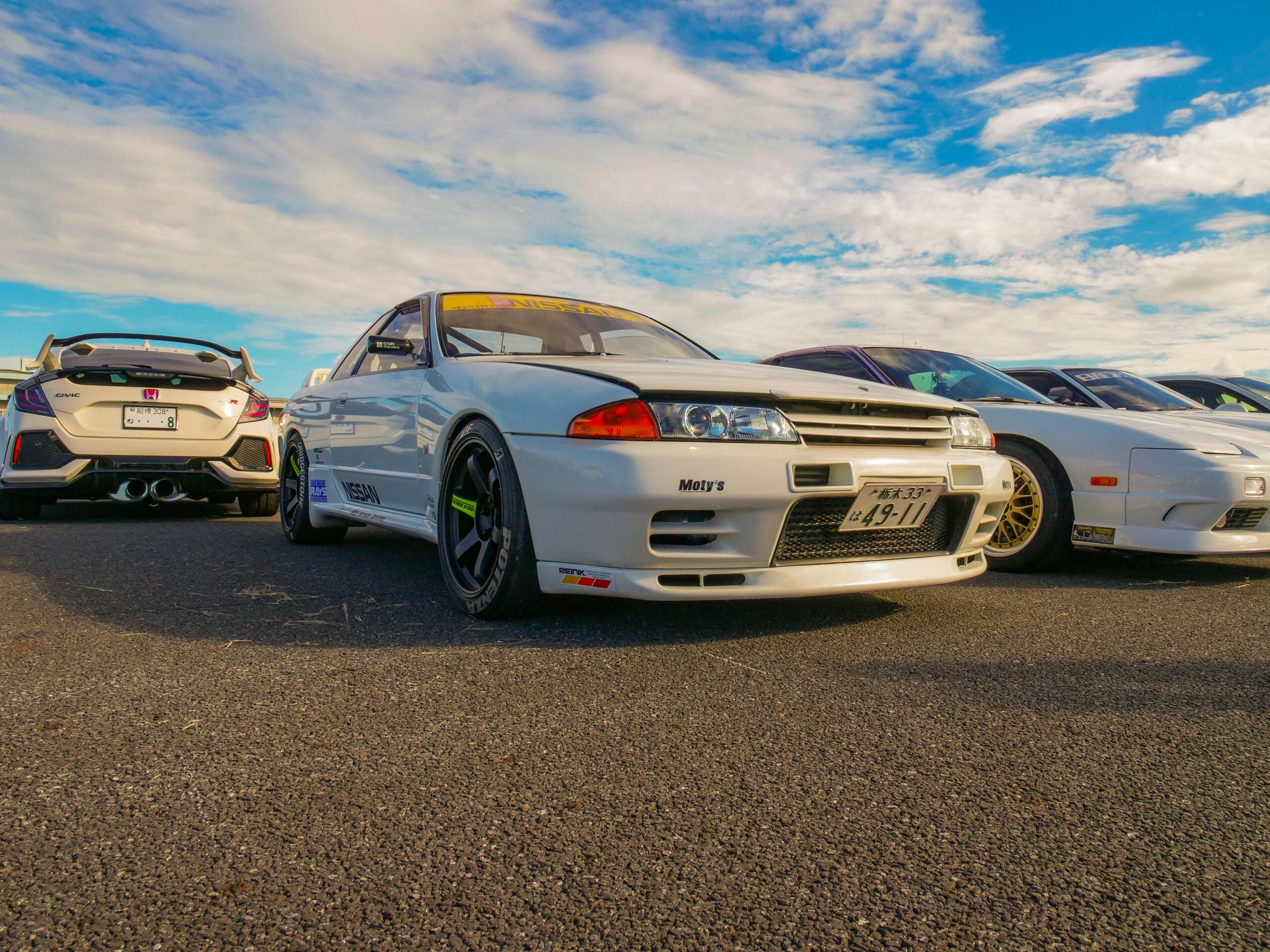A vibrant outdoor car meet showcasing iconic JDM cars including a Nissan Skyline and Honda Civic.