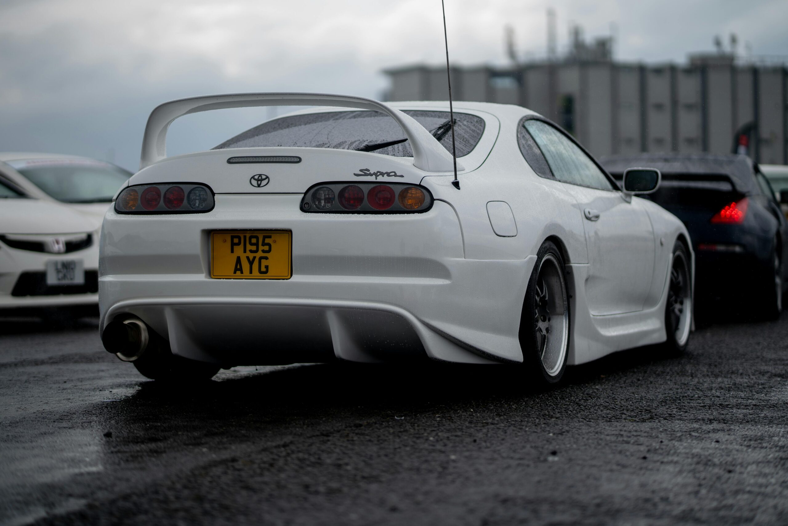 Rear view of a modified white Toyota Supra on a wet race track, showcasing its distinctive spoiler.