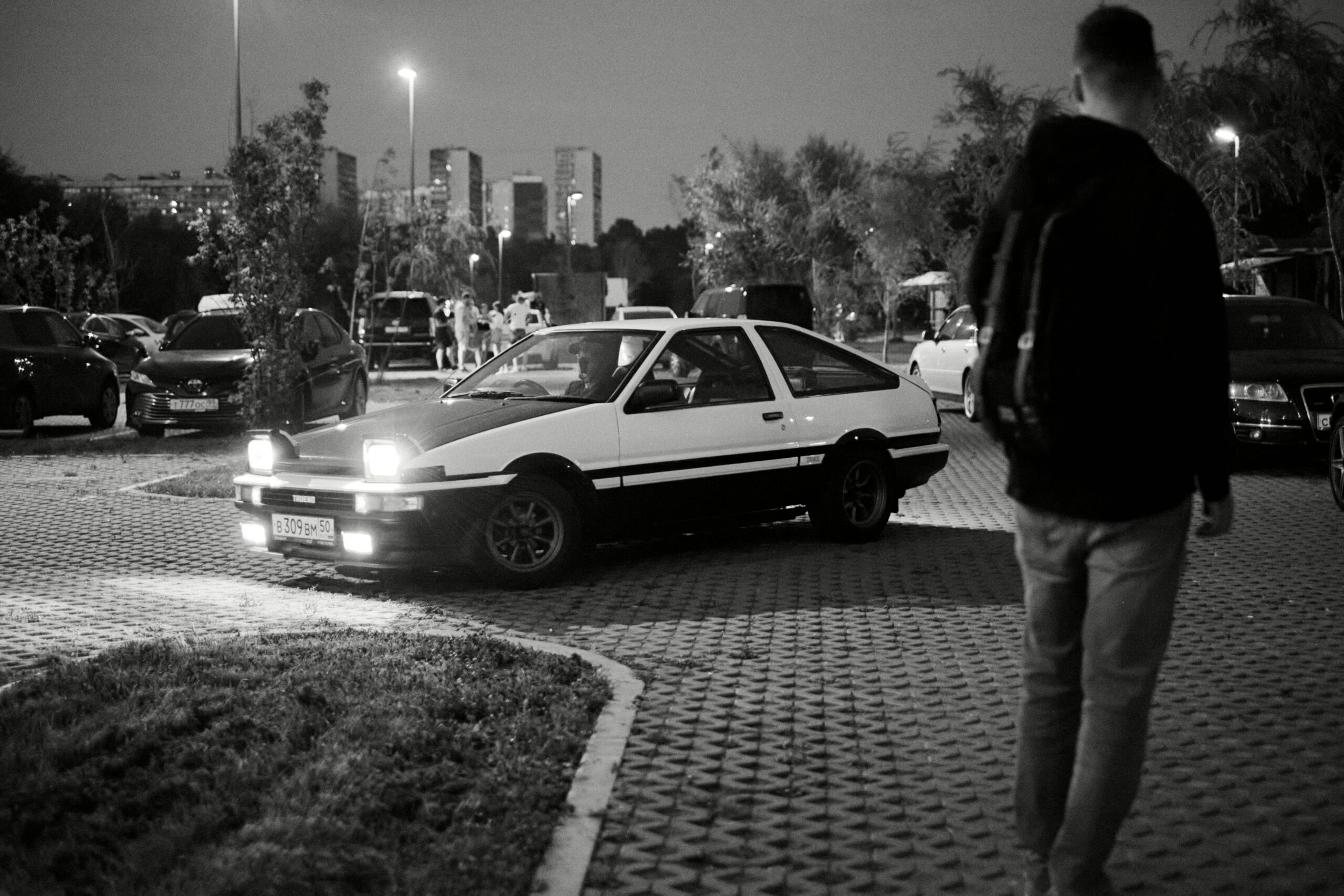 a man standing next to a parked car in a parking lot  By Jasper