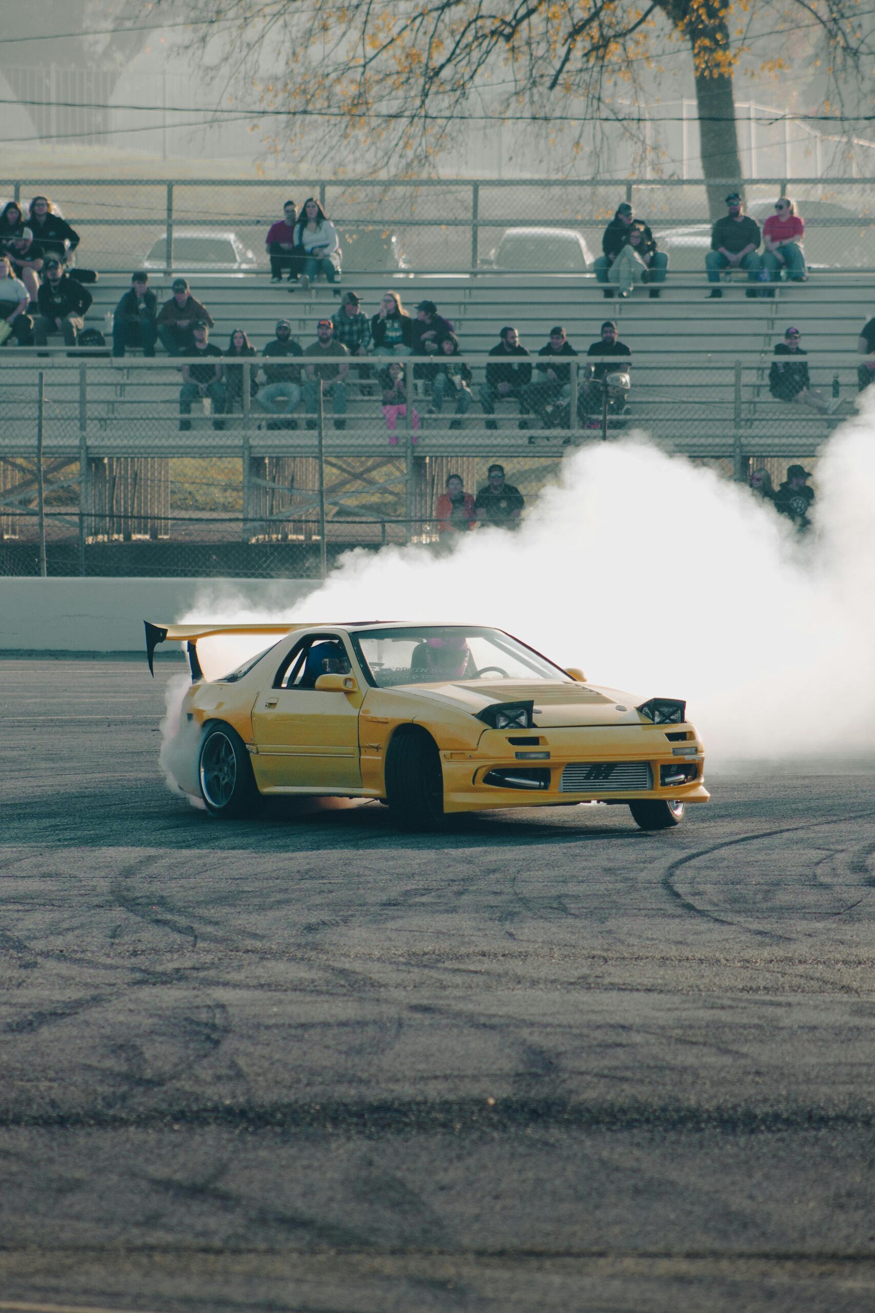 Yellow Mazda RX-7 drifts on track with spectators watching from stands.